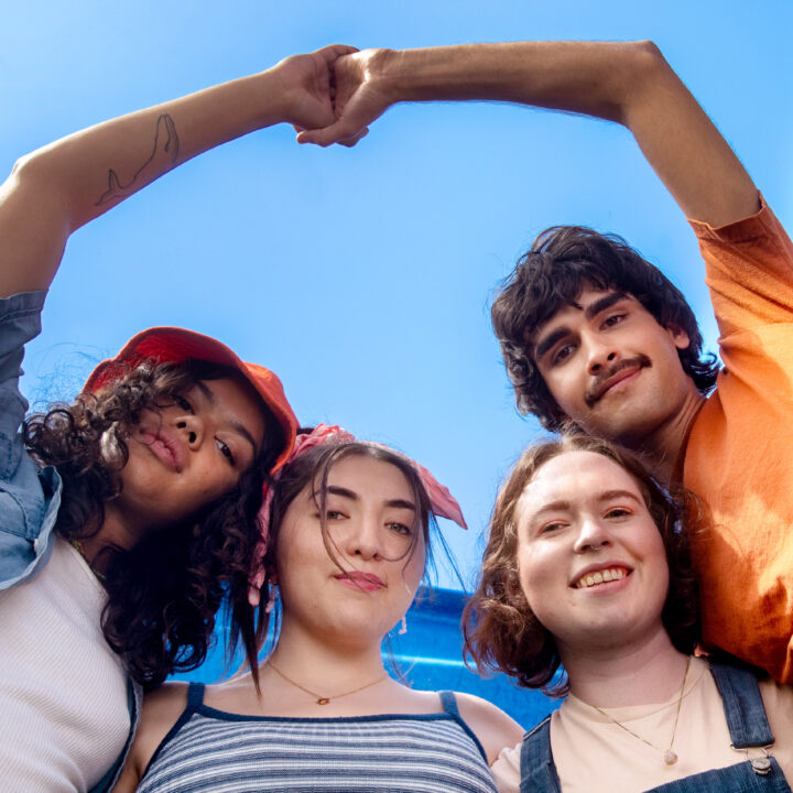 Group of LGBTQ youth, smiling and with their arms around each other, photographed from below with the sky behind them