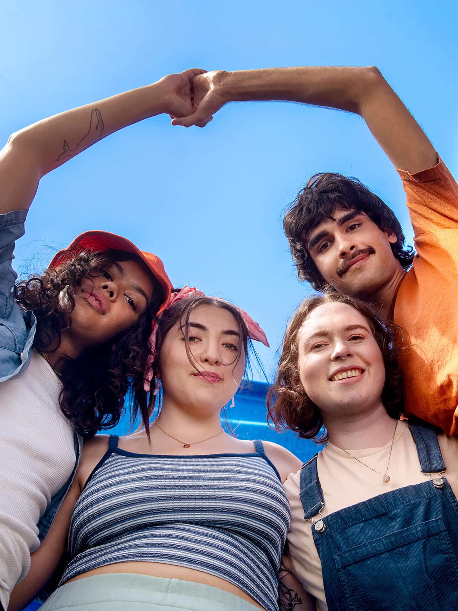 Group of LGBTQ youth, smiling and with their arms around each other, photographed from below with the sky behind them