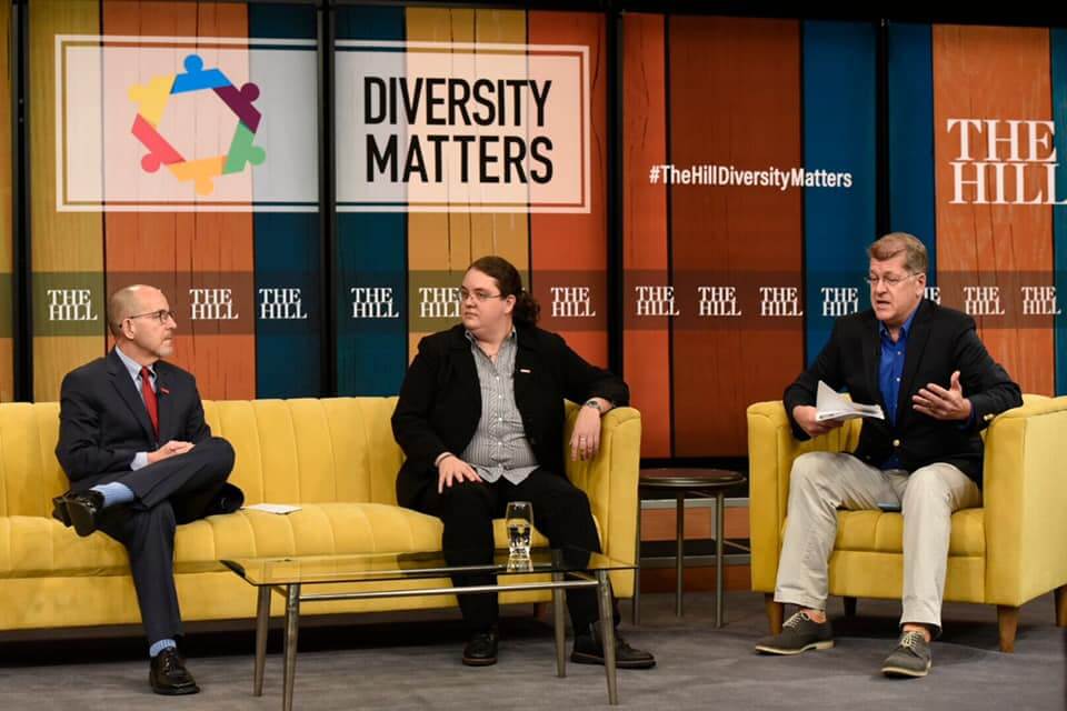 Casey sits on a yellow couch in conversation with two other people, beneath a Diversity Matters sign.