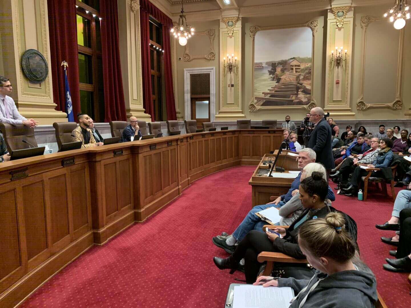 Troy stands at a podium in front of a small seated crowd, addressing a group of City Council members.