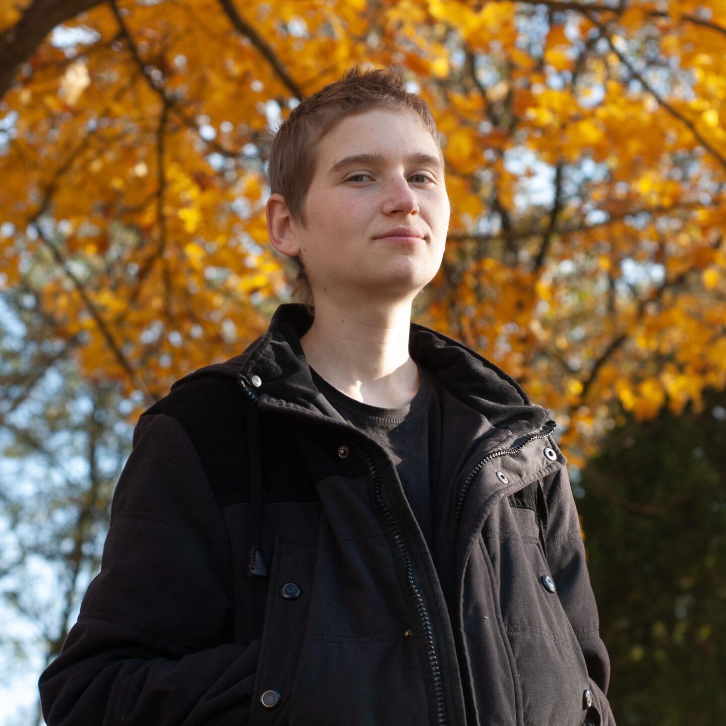 A young person in a large black coat standing outside on a colorful fall day.