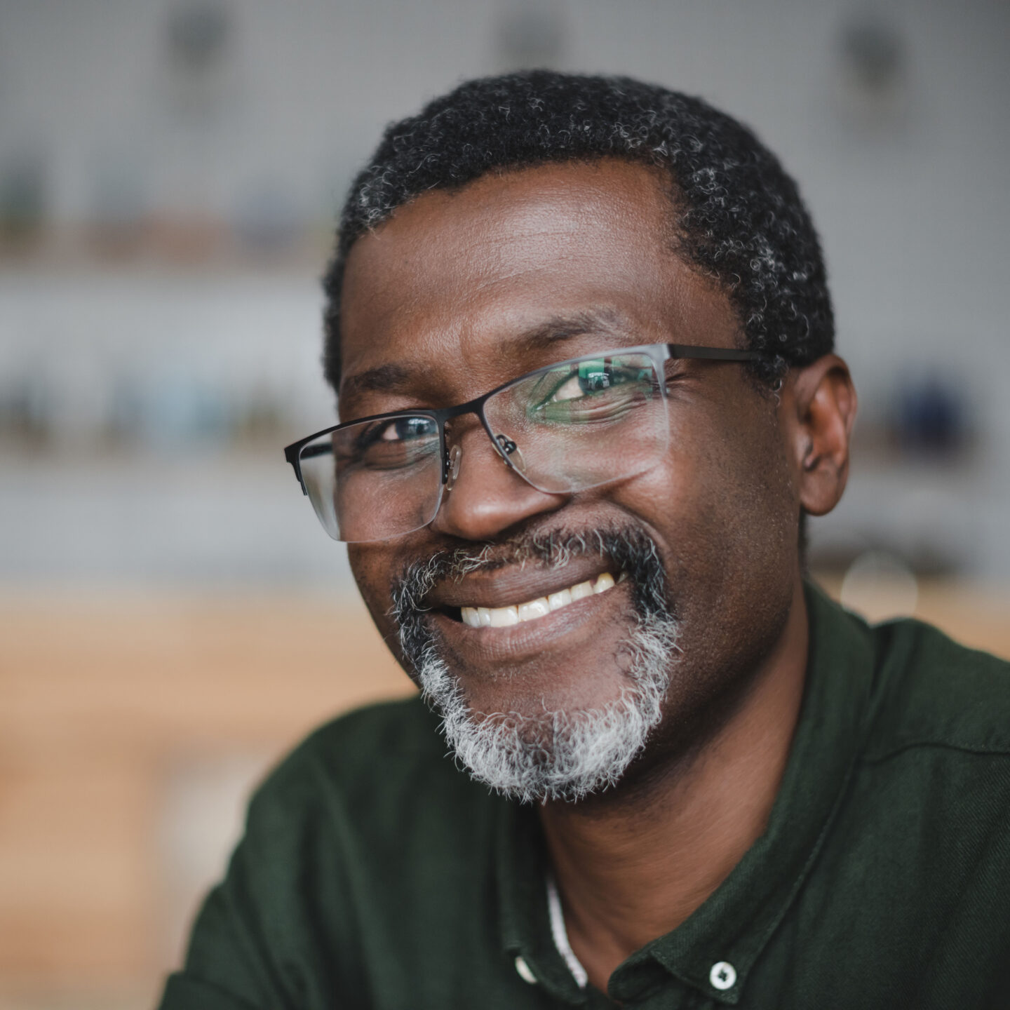 An adult with a grey beard and glasses smiling in their home.