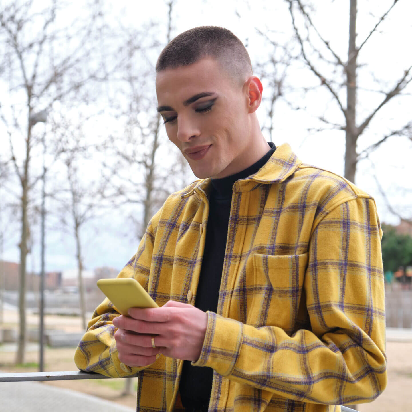 A person wearing a bright yellow flannel leaning on a balcony smiles while typing on their phone.