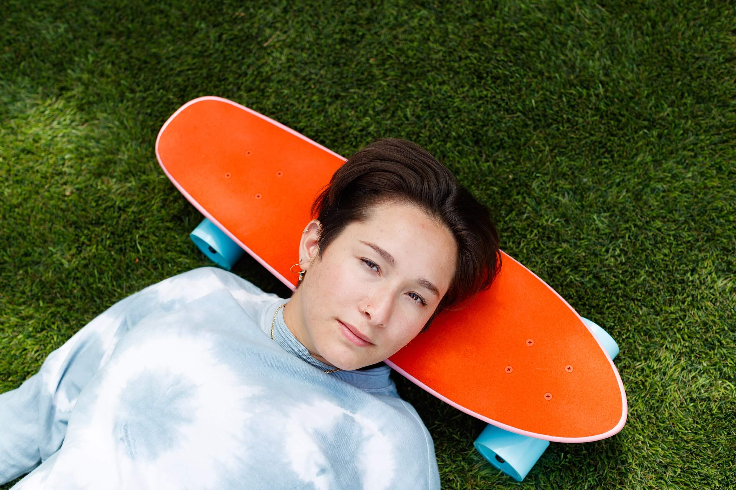 Portrait of a person laying on the grass, resting their head on an orange skateboard and looking into the camera