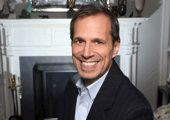 A smiling Jason Adams sitting in front of a fireplace.