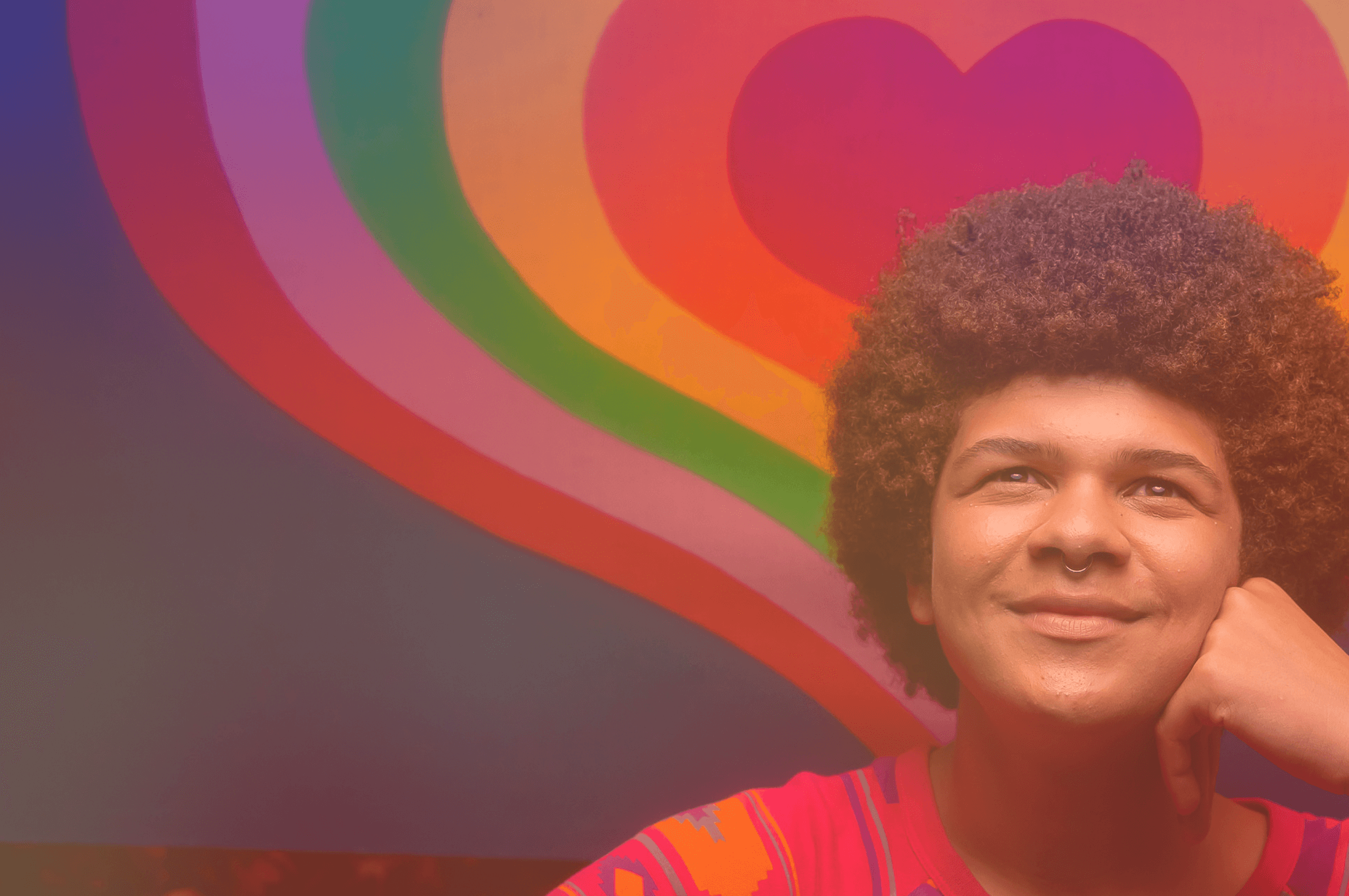 A photo of an LGBTQ+ young person smiling and looking up. They're posing in front of a rainbow heart background.