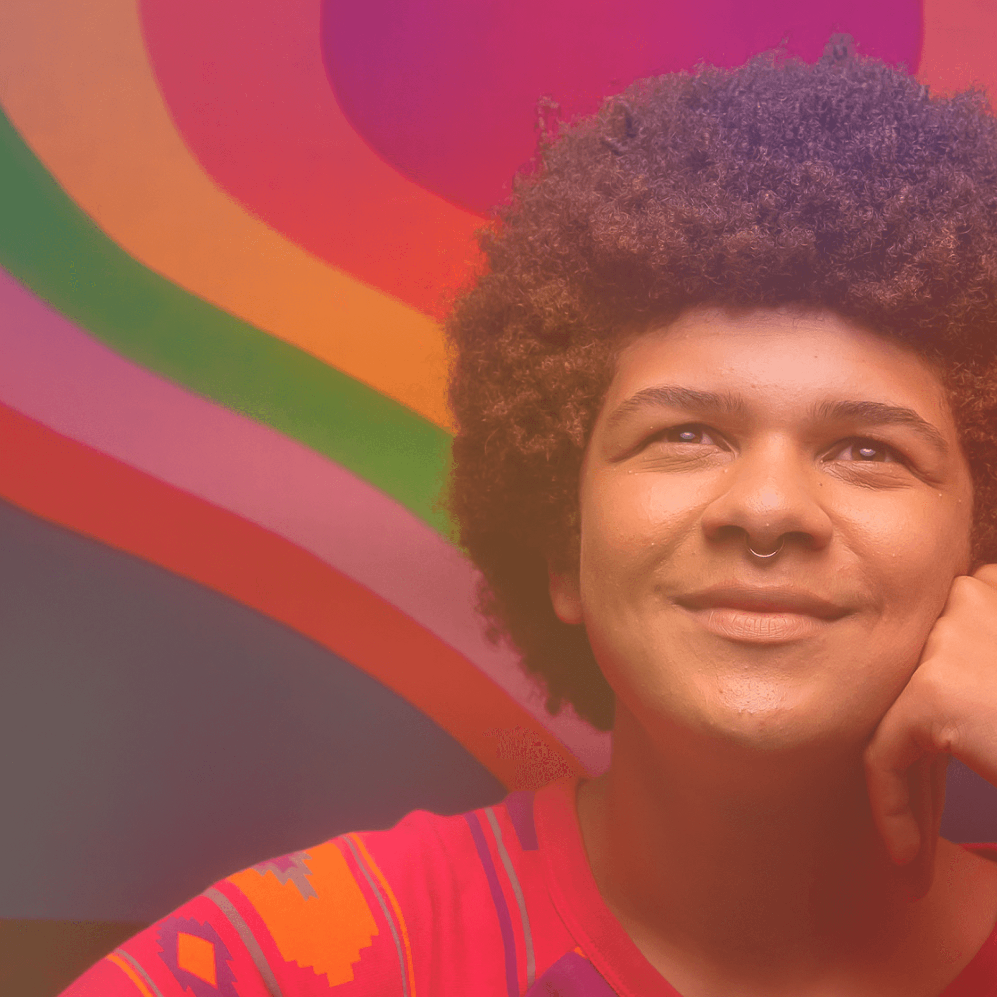 A photo of an LGBTQ+ young person smiling and looking up. They're posing in front of a rainbow heart background.